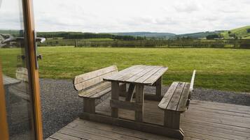 A wooden picnic table with benches on a wooden deck, overlooking a grassy field with a fence and distant hills.
