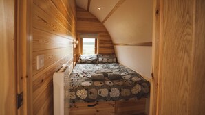 A narrow, wooden-paneled room with a sloped ceiling, a bed with patterned bedding, and a window providing a view of the outdoors. - Wooden Cosy Cabin with Hot Tub in Mid Wales (Llanbrynmair)