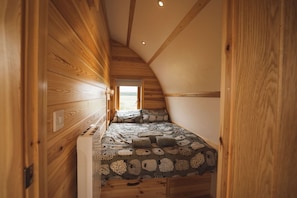 Een kleine kamer met houten lambrisering en een schuine kap, een bed met behang en een raam met uitzicht op het landschap.