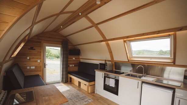 A compact kitchen with a built-in oven, microwave, and sink, wooden flooring, and a window overlooking a scenic landscape. - Wooden Cosy Cabin with Hot Tub in Mid Wales (Llanbrynmair)