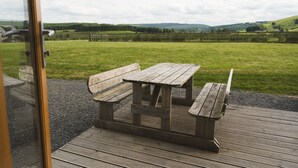 A wooden picnic table with benches on a wooden deck, overlooking a grassy field with a fence and distant hills. - Wooden Cosy Cabin with Hot Tub in Mid Wales (Llanbrynmair)