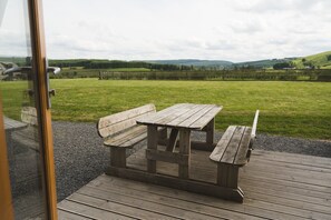 Een houten picknicktafel met bankjes op een houten terras, met uitzicht op een grasveld met een omheining en heuvels in de verte.