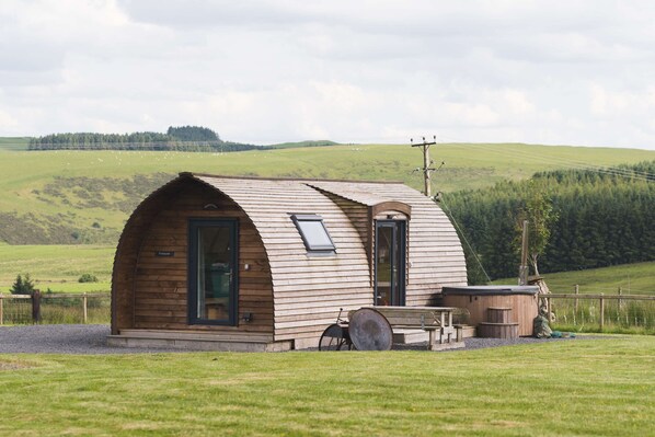 Een houten hut met een gebogen dak, een deur en een klein bijgebouwtje met een bankje en een tafel.