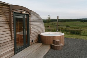 Eine Holzdiele mit Glastür, einem Whirlpool und Blick auf eine grüne Landschaft.