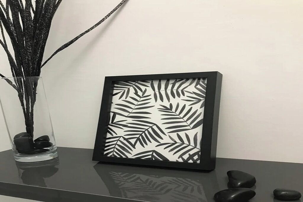 Een lijstwerk met een bladpatroon, een vaas met takken en enkele stenen op een reflecterend oppervlak.