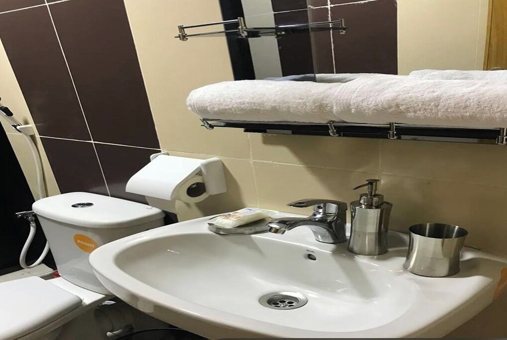 A bathroom with a white sink, a toilet, a towel rack with a white towel, a soap dispenser, and a metal cup.