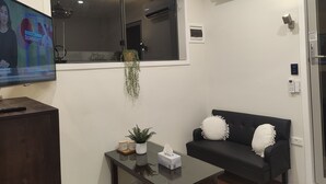 Uma sala de estar com televisão, um sofá preto, uma mesa pequena, uma janela e um aparelho de ar condicionado.