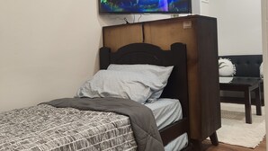 Quarto com uma cama, televisão na parede, cabeceira de madeira, sofá preto e parede branca.