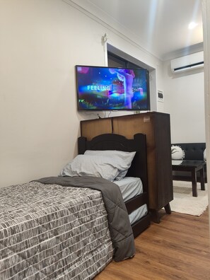 Ein Schlafzimmer mit einem Bett, einem an der Wand montierten Fernseher, einem Holz-Kopfteil, einem schwarzen Sofa und einer weißen Wand.