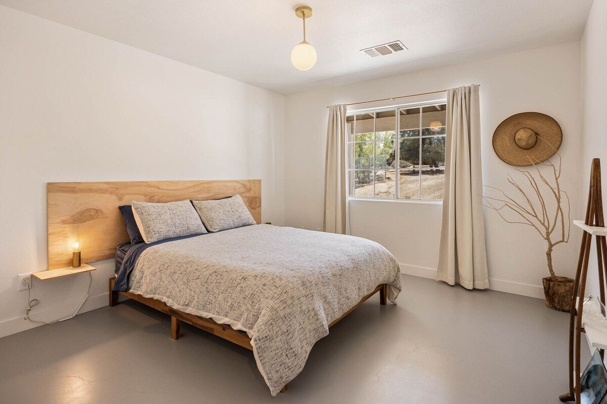 Een slaapkamer met een bed, nachtkastje, lamp, raam met gordijnen, en een hoed die aan de muur hangt.