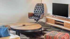 A modern living room with a round wooden table, a tufted leather chair, a flat-screen TV, and a large window with a view.