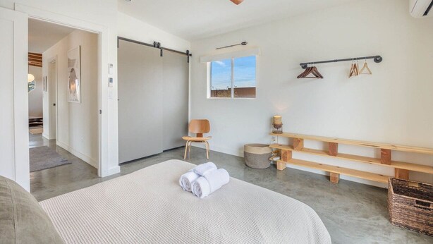 A bedroom with a bed, a ceiling fan, a window, a bench, and a wall-mounted rack with clothes.