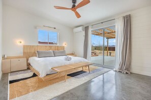 Un dormitorio moderno con cama, mesitas de noche, ventilador de techo y un amplio ventanal corredero que da a un espacio exterior.