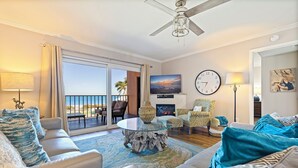 A spacious living room with a glass coffee table, a ceiling fan, a large TV, and a balcony with a view of the ocean. - Reef Club 209 - Oceanview Retreat w/ Pool, Tennis & Sunset Balcony (Indian Rocks Beach)