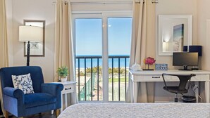 A hotel room with a bed, a desk with a computer, a chair, a lamp, and a view of the sea through a sliding door. - Reef Club 209 - Oceanview Retreat w/ Pool, Tennis & Sunset Balcony (Indian Rocks Beach)