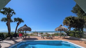 A pool area with a view of the beach, palm trees, and thatched umbrellas.