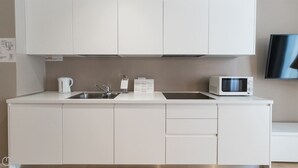 A modern kitchen with white cabinetry, a stainless steel sink, a kettle, a microwave, and a flat-screen TV mounted on the wall.