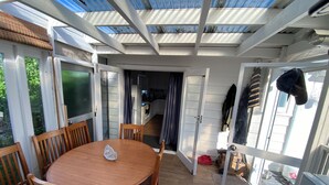 A covered patio with a wooden table and chairs, a kitchen area in the background, and a view of the outside.