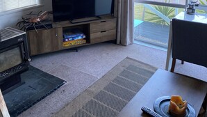 A living room with a flat-screen TV, wooden entertainment unit, and a view of the outdoors through large windows.