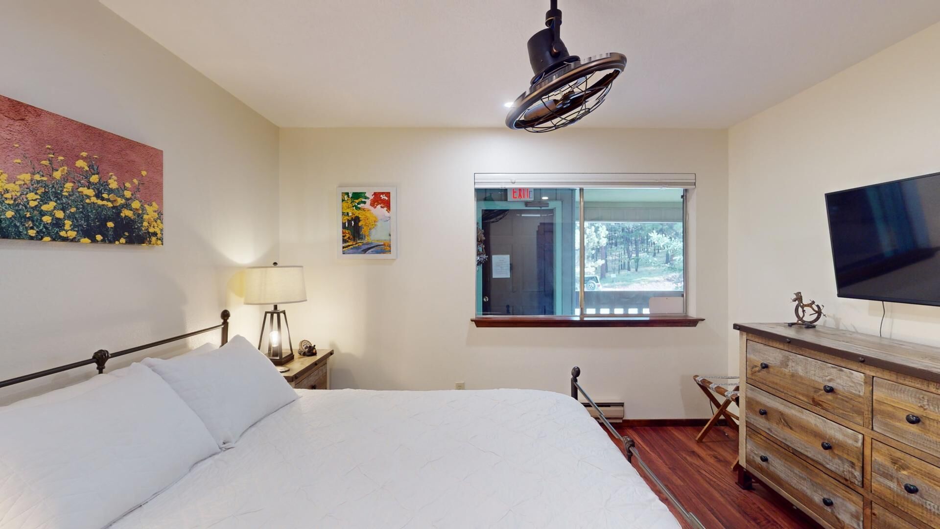 Een slaapkamer met een bed, nachtkastje, lamp, dressoir, televisie en raam met uitzicht op bomen.