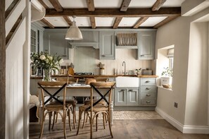 A kitchen with wooden beams, light blue cabinets, a wooden dining table with chairs, and a hanging light fixture. - Dog Friendly Cotswold Holiday Cottage - The Forge (Upper Oddington)