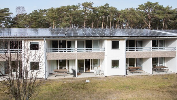 Un bâtiment à deux étages, doté d’un toit plat recouvert de panneaux solaires, d’un balcon à l’étage supérieur et d’un petit espace extérieur aménagé avec des bancs.