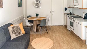 Un espace de vie compact comprenant un canapé gris, une table basse ronde en bois, une table à manger avec des chaises et une kitchenette équipée d’une cuisinière et d’un évier.