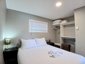 A neatly made bed with pillows, a bedside lamp, a window with blinds, and a closet with hanging clothes.