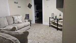 A living room with a beige carpet, a grey sofa, and a small side table with a lamp. - Charming 2-bedroom house in close proximity to the YUM Ctr, the Downs, etc. (Louisville)