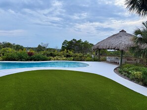 A swimming pool with a thatched-roof pavilion, surrounded by well-maintained grass and tropical plants. - Indian River Retreat  - Main House and Cottage (Jensen Beach)