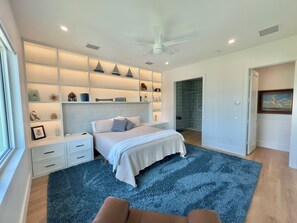 A modern bedroom with a bed, a nightstand, a built-in shelf with decorative items, a shower, and a painting on the wall. - Indian River Retreat  - Main House and Cottage (Jensen Beach)