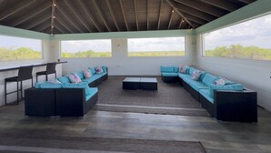A covered outdoor seating area with a long sofa and chairs, a small table, and large windows offering views of the landscape.