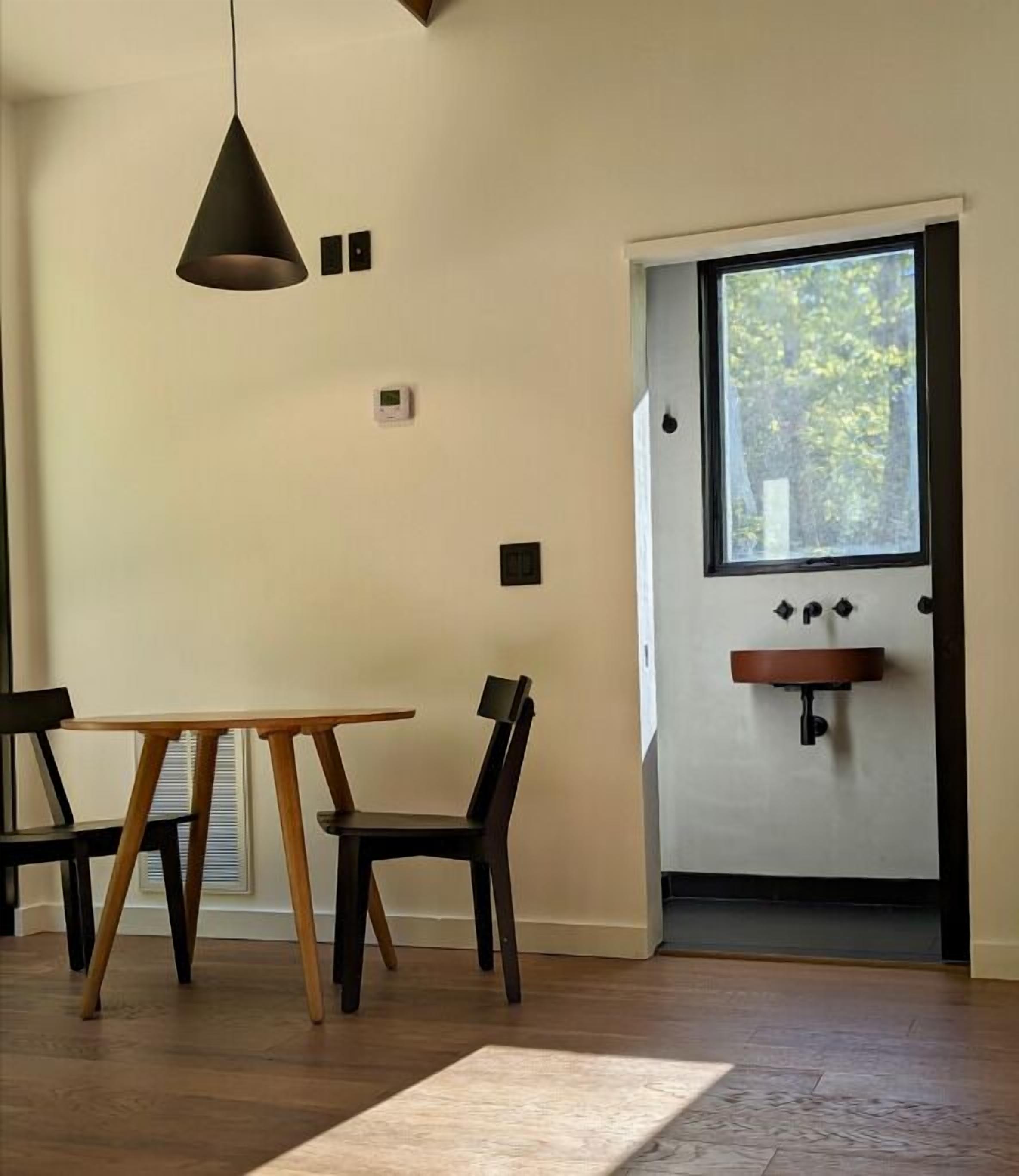 A dining area with a wooden table and chairs, a hanging light fixture, a window with a view of trees, and a built-in shelf with a sink.