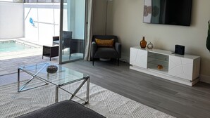 A modern living room with a glass coffee table, a grey sofa, a white console table with decorative items, a floor lamp, and a TV mounted on the wall. - 3-bedroom house in Orlando close to Disney with Customizable children's room (Clermont)