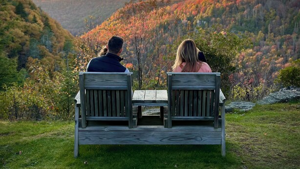 Deux personnes sont assises sur un banc en bois, face à un paysage pittoresque d’une vallée boisée.