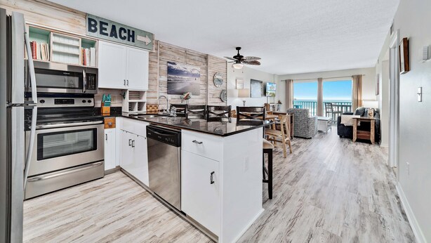 Een moderne keuken met witte kasten, een ingebouwde oven en een magnetron. Er is een eethoek met een tafel en stoelen, en uitzicht op het strand door grote ramen.