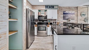 Een moderne keuken met een roestvrijstalen koelkast, magnetron en oven, houten kastjes en een bord met de tekst 'BEACH' aan de muur.