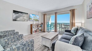 Een woonkamer met een bank, een salontafel en een boekenkast. Er is een balkon met uitzicht op het strand.