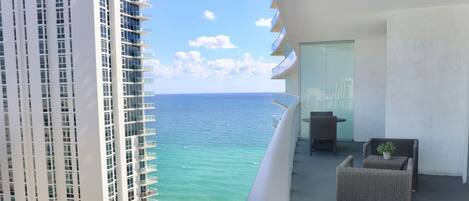 A balcony with a view of the ocean, a high-rise building, and outdoor furniture.
