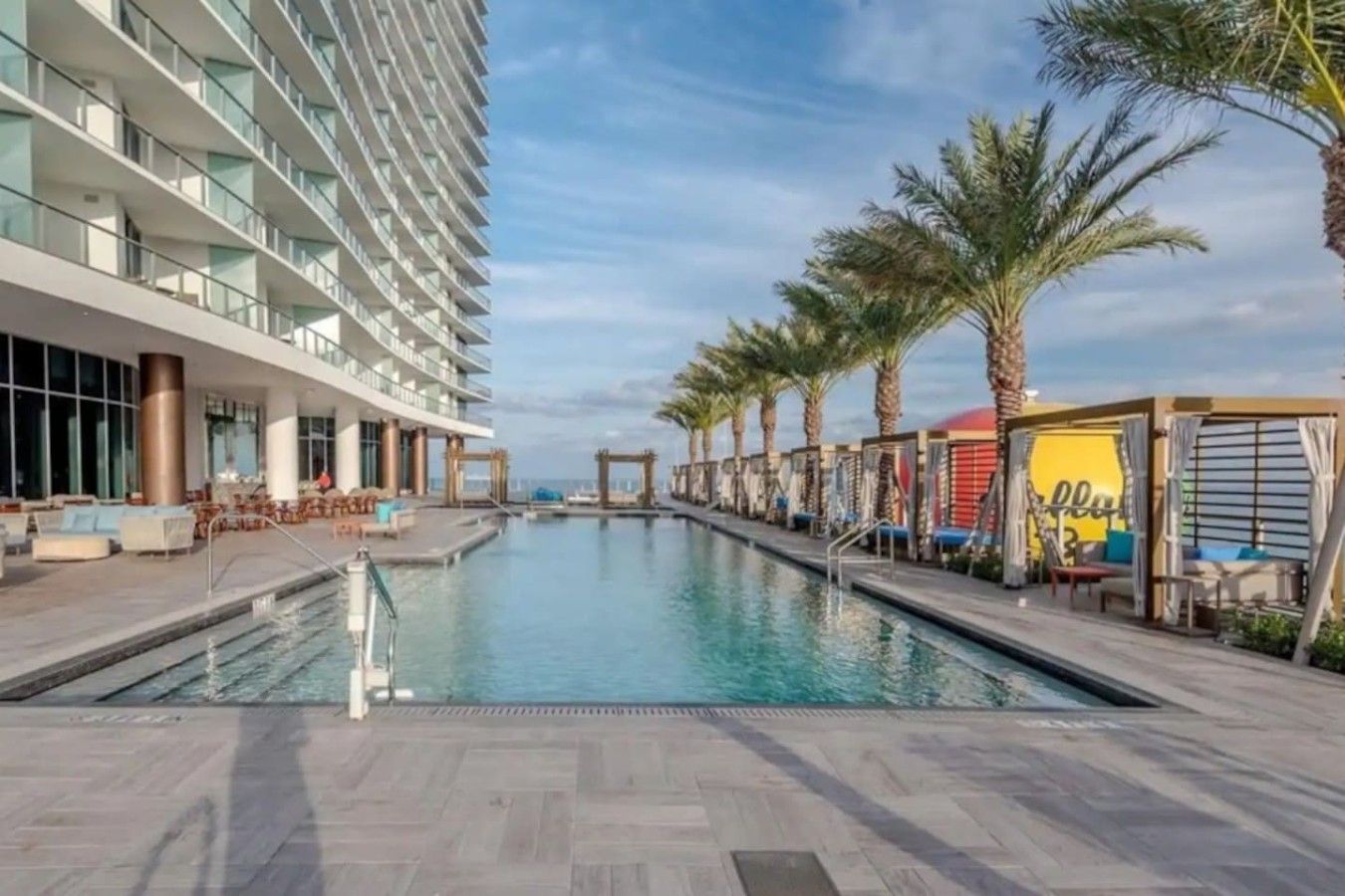A swimming pool with lounge chairs and palm trees, surrounded by a modern building with balconies.