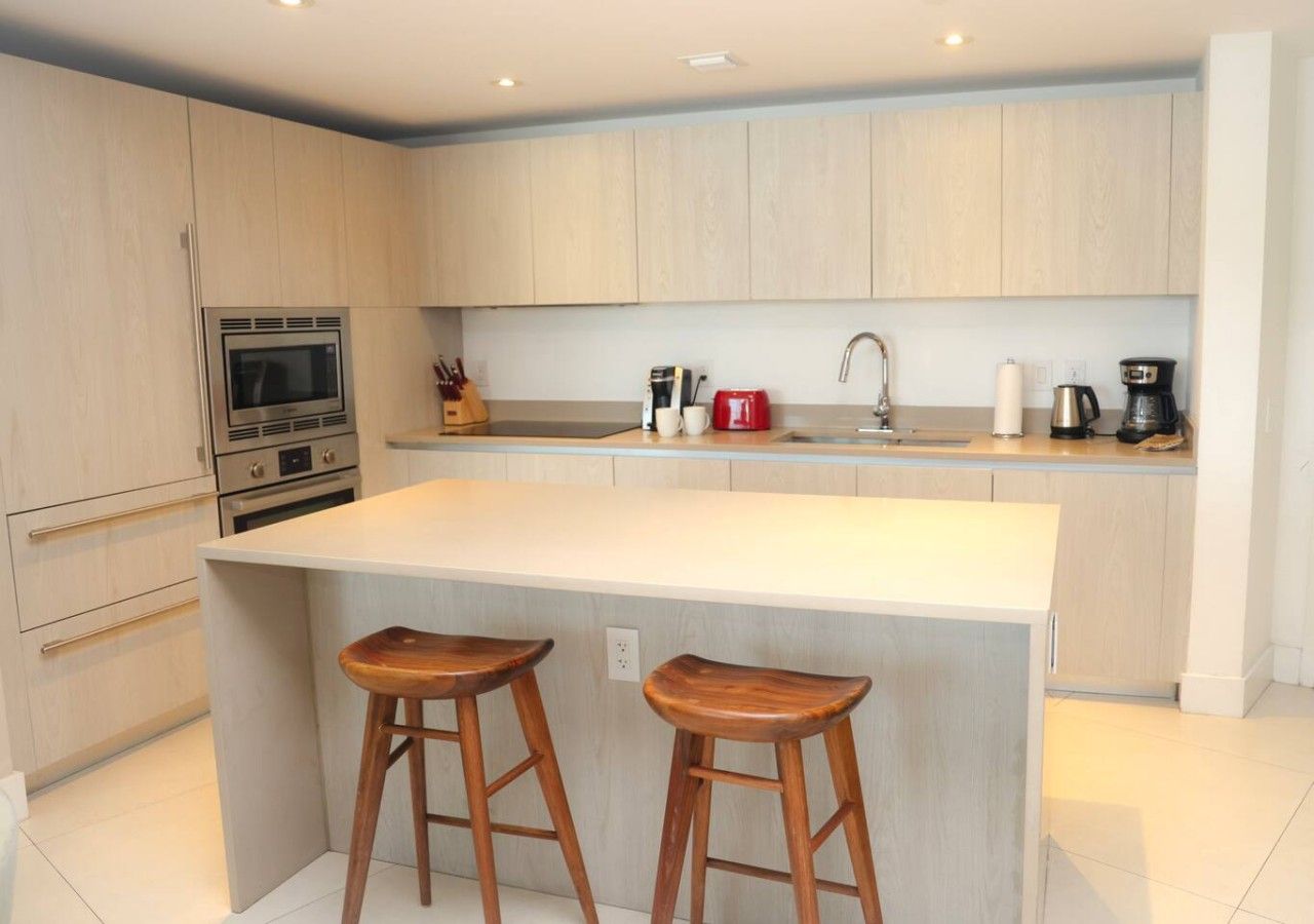 A modern kitchen with a central island, two wooden stools, stainless steel appliances, and a light-colored countertop.