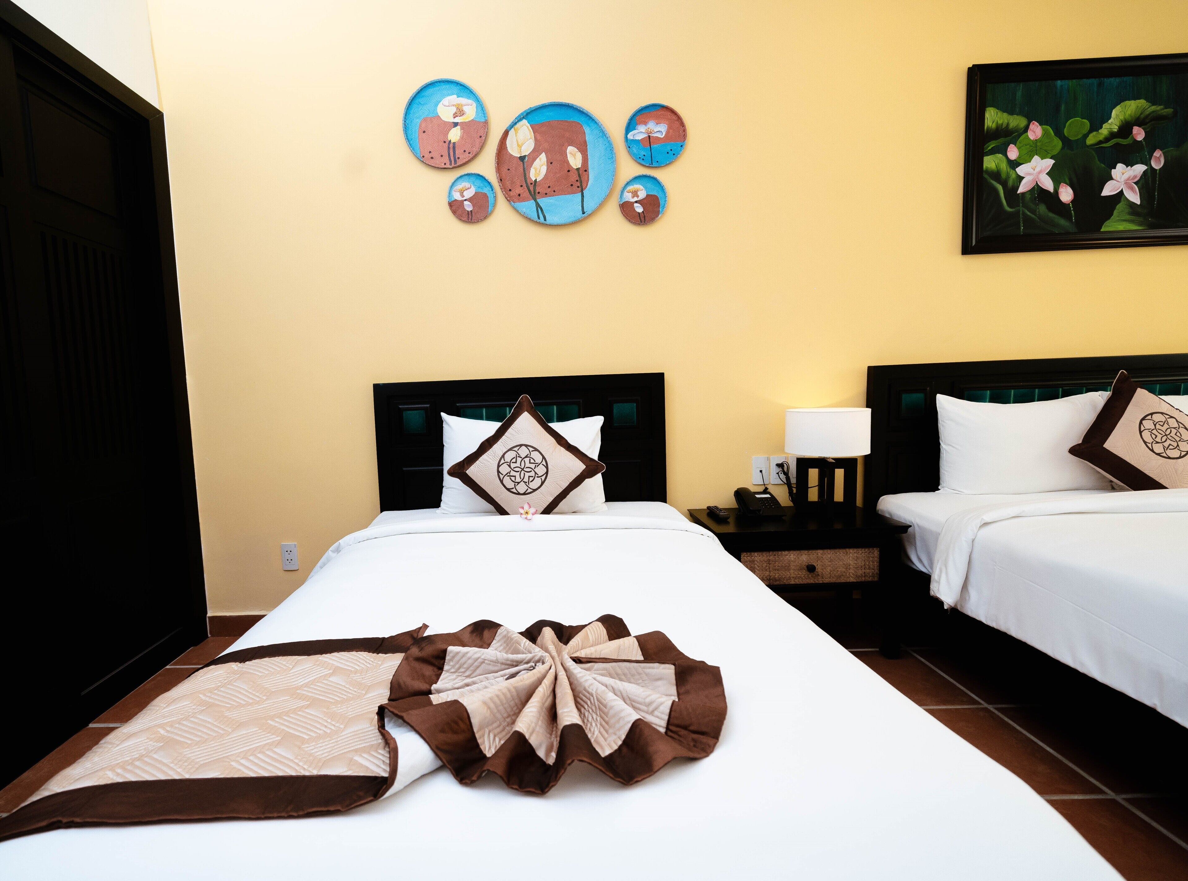 A hotel room with two beds, a nightstand with a lamp, a painting on the wall, and a decorative pillow on the headboard.