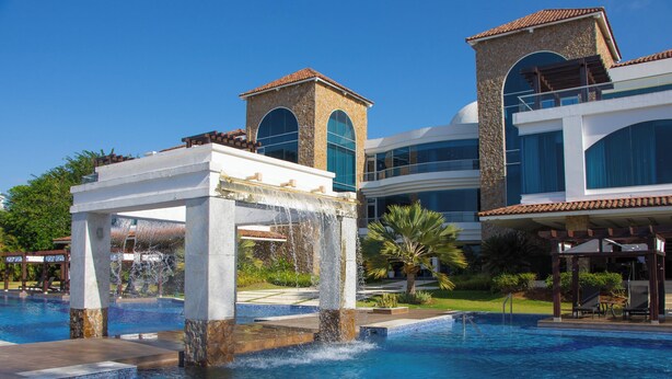 Uma área de piscina com um elemento aquático e um edifício com janelas em arco e uma torre.