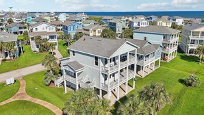 A multi-story residential building with a balcony on the second floor, surrounded by a well-maintained lawn and palm trees. - Mermaid's Cove -Beachside Bliss Pointe West Home, Reduced Rate! (Galveston)