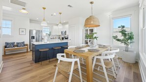 A modern kitchen with white cabinets, a central island, and bar stools. There is a dining table with placemats and chairs. - Mermaid's Cove -Beachside Bliss Pointe West Home, Reduced Rate! (Galveston)