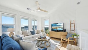 A modern living room with a white sofa, a blue sofa, a round coffee table, a television, and a view of a cityscape through large windows. - Mermaid's Cove -Beachside Bliss Pointe West Home, Reduced Rate! (Galveston)