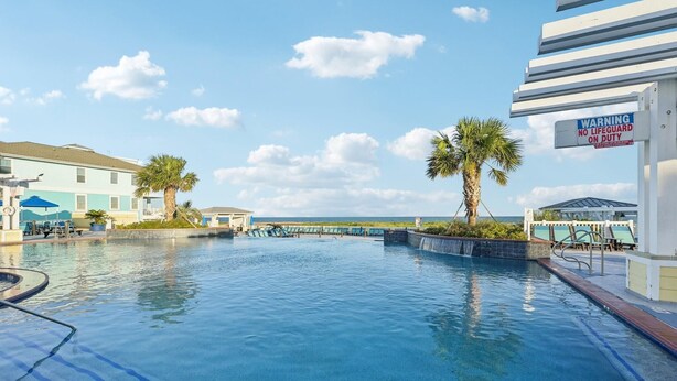 A swimming pool with lounge chairs and palm trees, surrounded by buildings. - Mermaid's Cove -Beachside Bliss Pointe West Home, Reduced Rate! (Galveston)