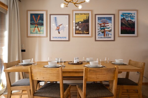 Un comedor con una mesa de madera dispuesta para seis personas, carteles enmarcados en la pared y una ventana con cortinas.