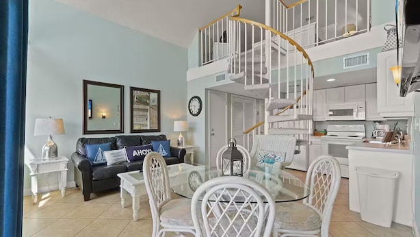 A modern living room with a glass dining table, white wicker chairs, a black leather sofa with blue cushions, a white coffee table, and a spiral staircase leading to a loft bedroom. - Welcome to SEA-Nanigans. Unit #1366. 
A charming condo with Gulf Views. (Gulf Shores)