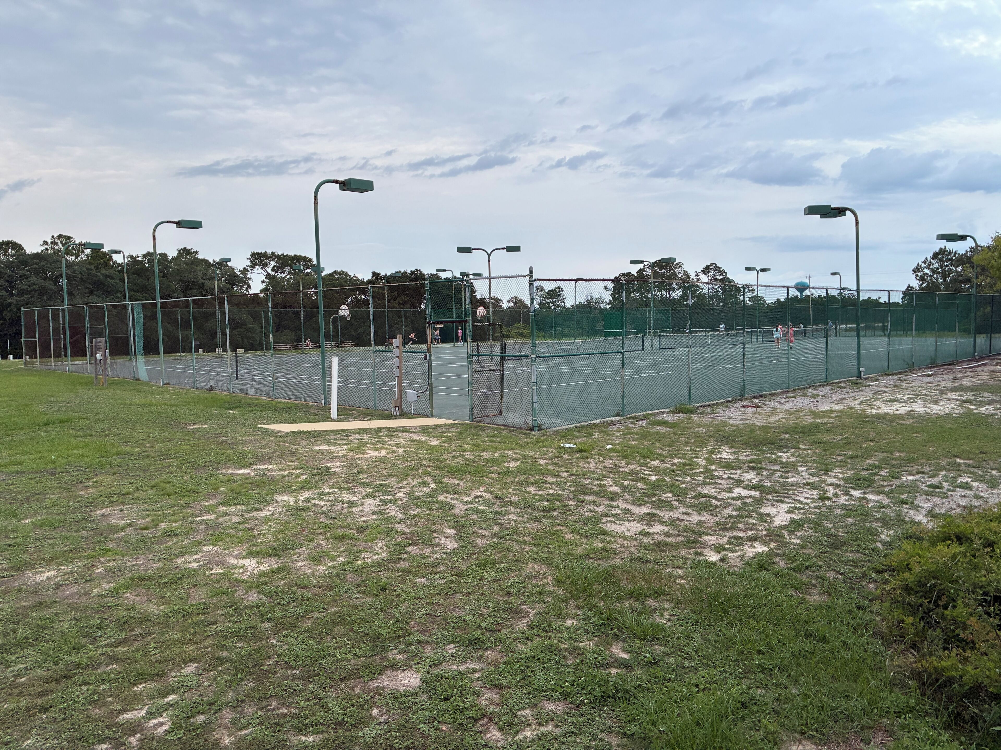 An outdoor tennis court with a fence, street lamps, and trees in the background.